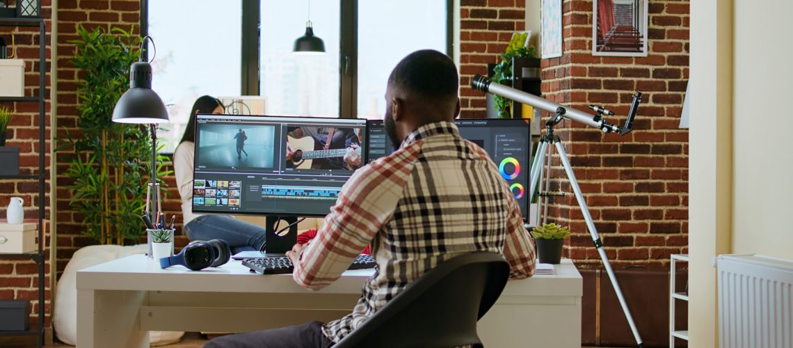 African american freelancer editing a background video at his home office, mixing clips on dual monitors. Professional man employs tech tools for creative and multimedia projects. Camera A.