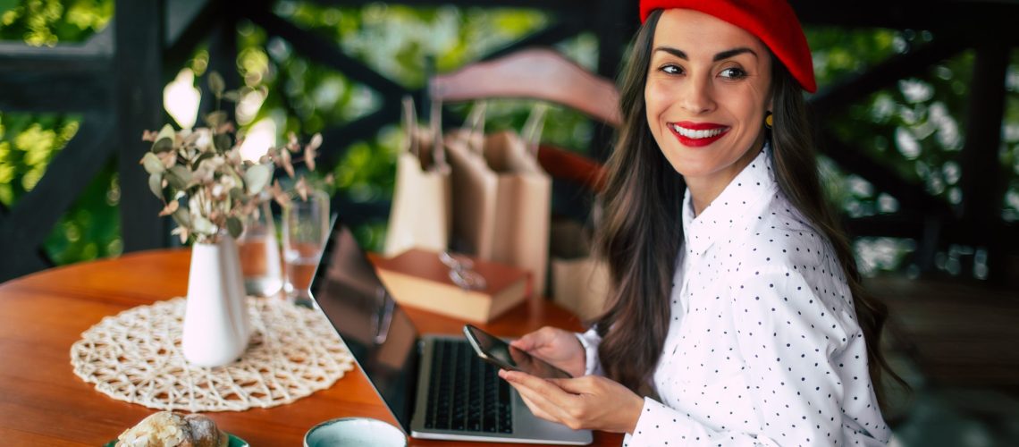 Gorgeous smiling cheerful brunette woman in a red beret resting and having breakfast in city cafe sitting at table on the terrace and using her smart phone for shopping online or chatting