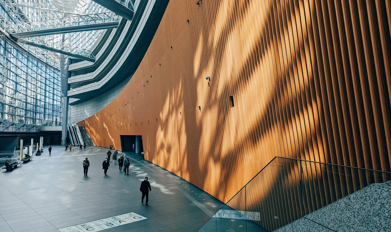 Passants dans le vaste hall d’un centre de congrès, sous une verrière, le long d’un mur courbe en lames de bois